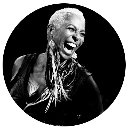 Black and white photo of a woman with short hair laughing joyfully. She is wearing large feather earrings and a sleeveless top. The background is dark, emphasizing her expressive smile and lively demeanor.