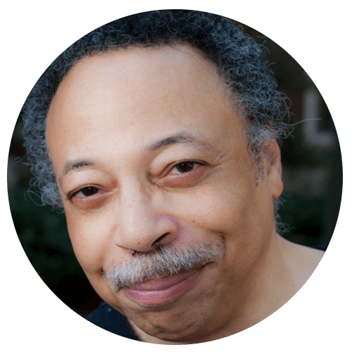 A middle-aged man with short curly gray hair and a mustache smiles softly at the camera. He is wearing a dark shirt and is outdoors with blurred greenery in the background.