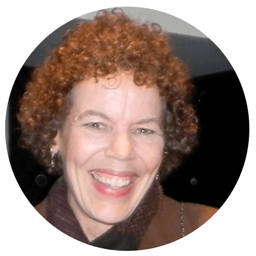 A person with short, curly hair smiles widely at the camera. They are wearing a dark brown coat and a burgundy scarf. The background is indoors with dark and light areas.