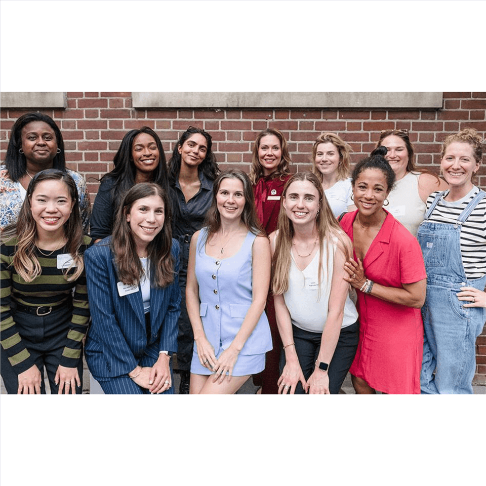 A group of thirteen women smiling and posing together in front of a brick wall. Most are dressed in business casual or casual attire, with a mix of dresses, blouses, and blazers.