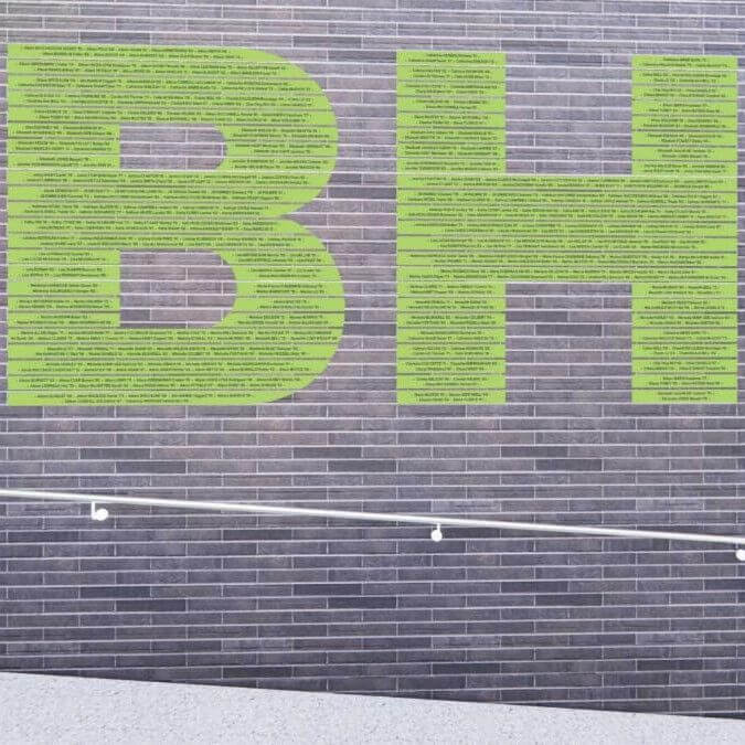 Large green letters "BH" are displayed on a gray brick wall. The surface in front of the wall is pale, and a white handrail runs diagonally across the lower part of the image.