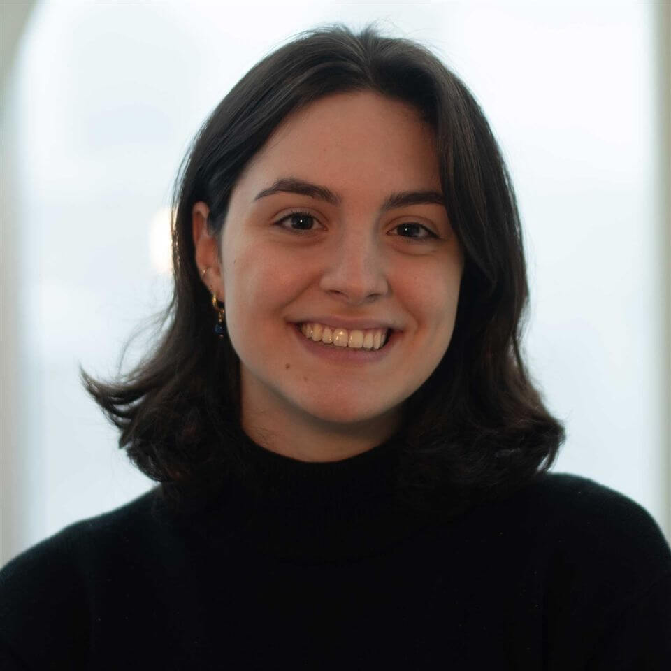 A young woman with shoulder-length dark brown hair, wearing a black turtleneck sweater, smiles at the camera in a softly lit indoor setting with a blurred background.