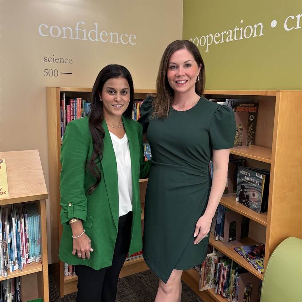 Two women stand smiling in front of bookshelves in a library. Both wear green outfits. Words like “confidence” and “cooperation” are visible on the wall behind them, along with shelves filled with books and colorful book spines.