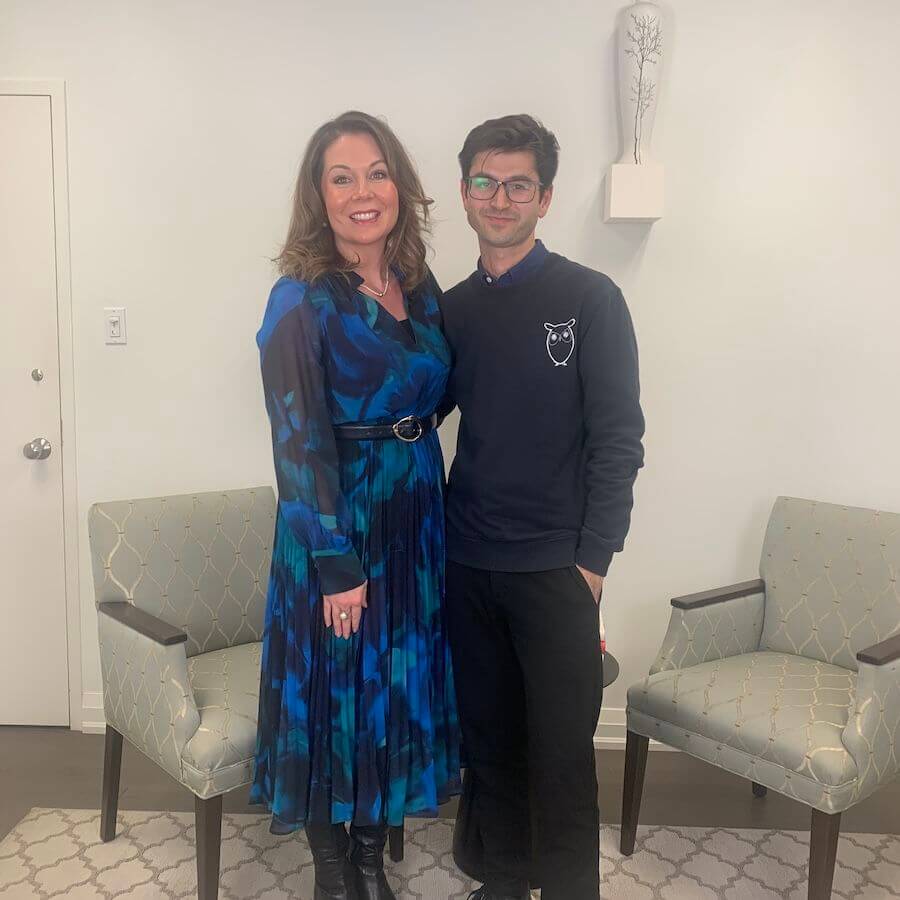 Two people stand side by side, smiling at the camera in a room with light walls and two patterned chairs. The woman wears a blue and black dress; the man wears glasses and a black sweater with an owl design.
