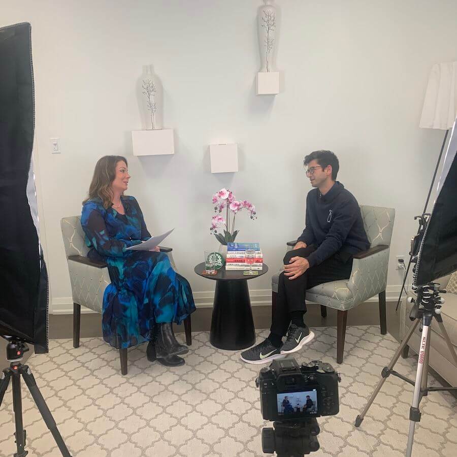 A woman in a blue dress interviews a man in dark clothing. They sit on armchairs with a small black table between them, holding books and flowers. Professional lights and a camera are set up in the room.