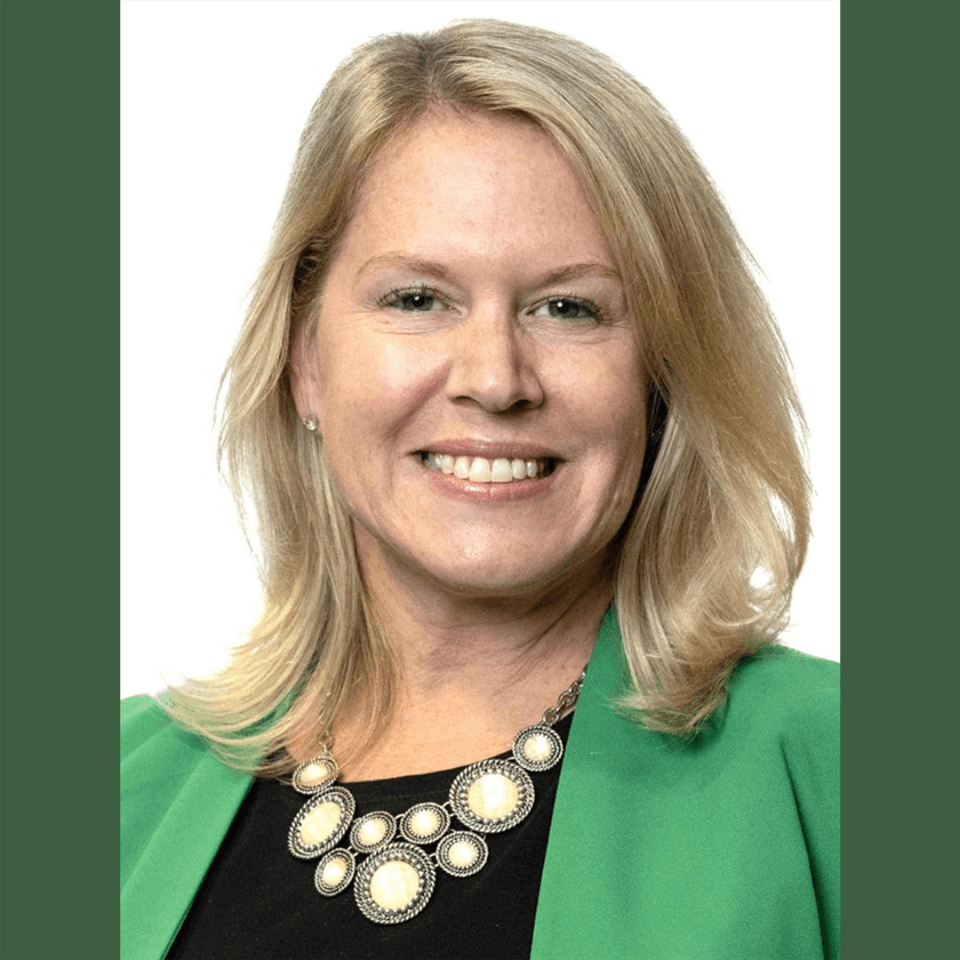 A woman with blonde hair, wearing a green blazer and a black top with a statement necklace, smiles in front of a white background.