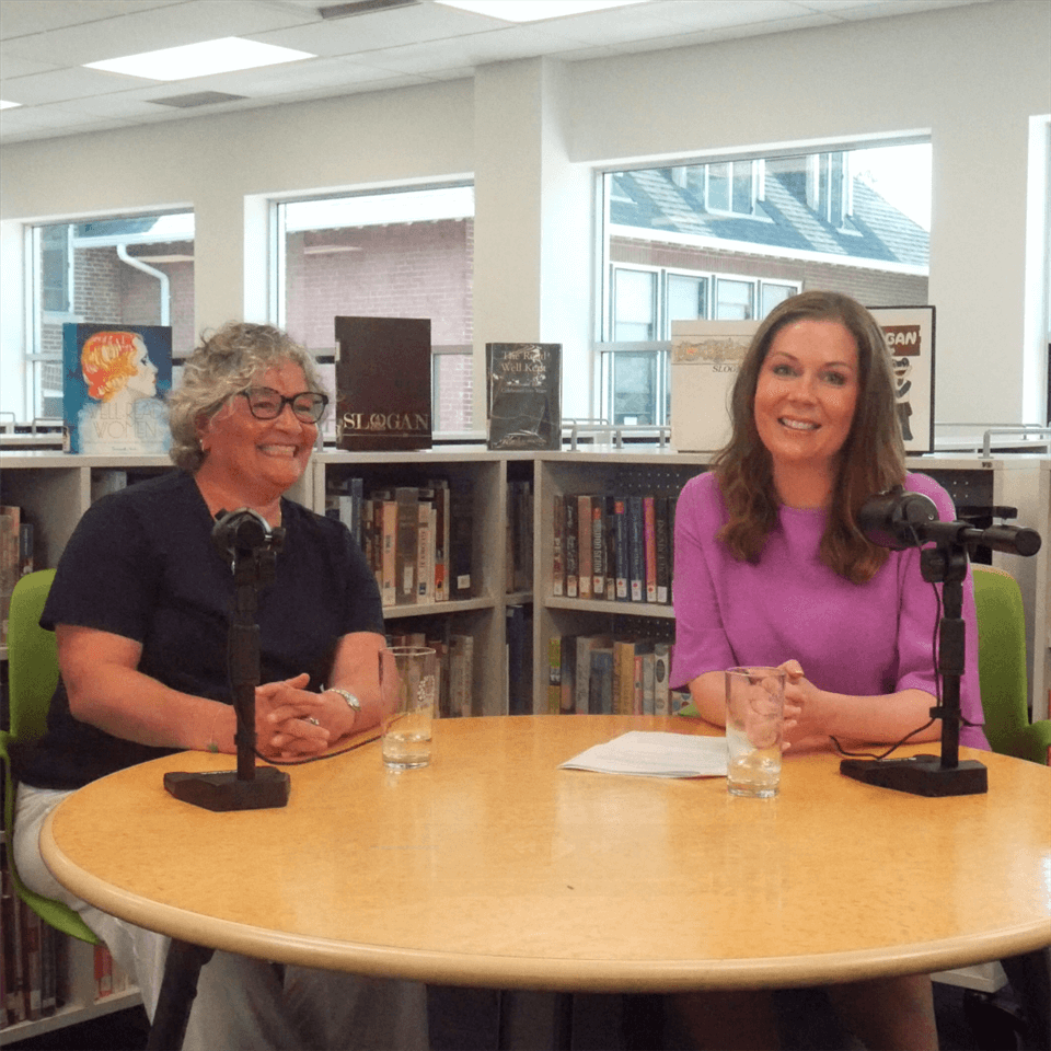 Two women sit at a round table with microphones and glasses of water in a bright library setting. Bookshelves and large windows are visible in the background. Both women are smiling.