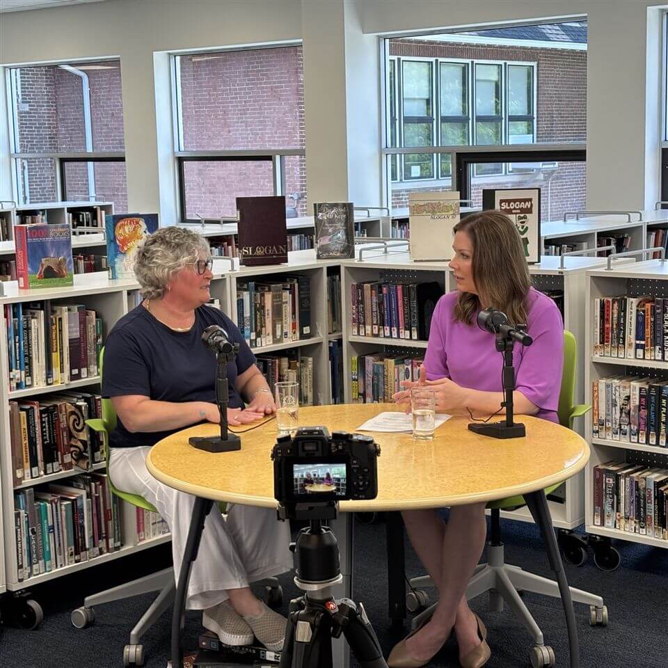 Two women sit at a round table with microphones and glasses of water, having a conversation in a library. Bookshelves and windows are in the background, and a camera on a tripod is recording them.