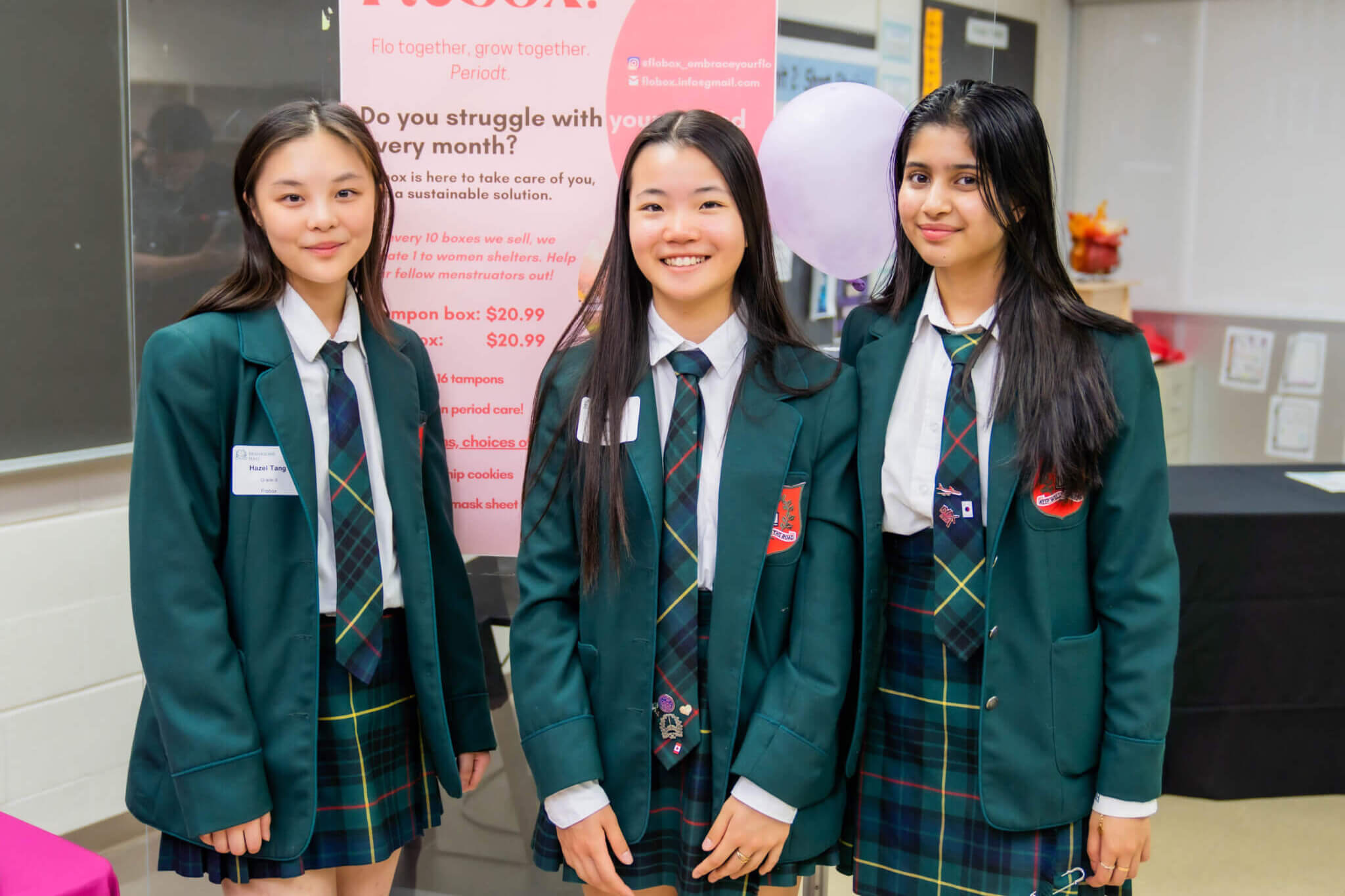 Three female students in green school uniforms stand indoors, smiling at the camera. Behind them is a pink informational poster and a table with promotional materials.