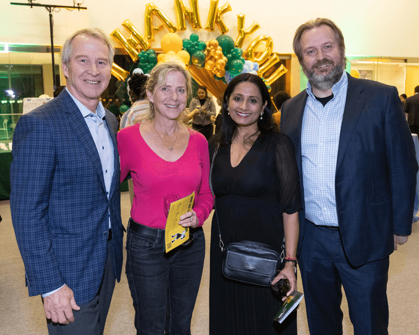 Four adults pose together at an indoor event in front of gold "THANK YOU" balloons and green and yellow decorations. Two women stand between two men; one holds a glass of wine and another holds a yellow booklet.