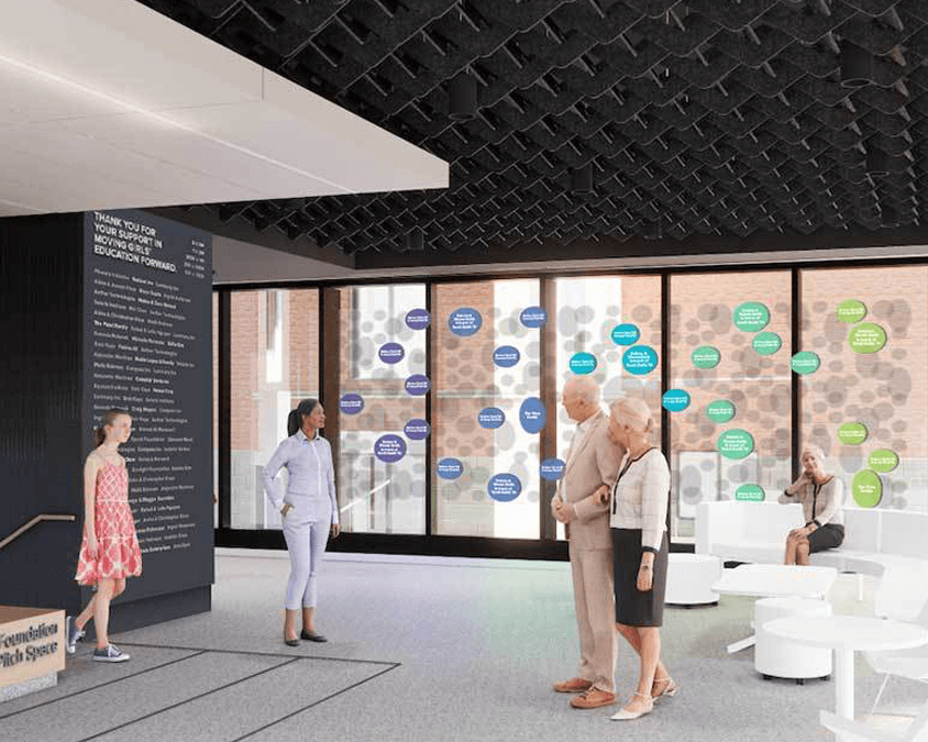 A group of people stands and sits in a modern, bright room with large windows displaying colorful decals. The space has minimalistic furniture and a wall of donor names. One person reads a sign near the entrance.