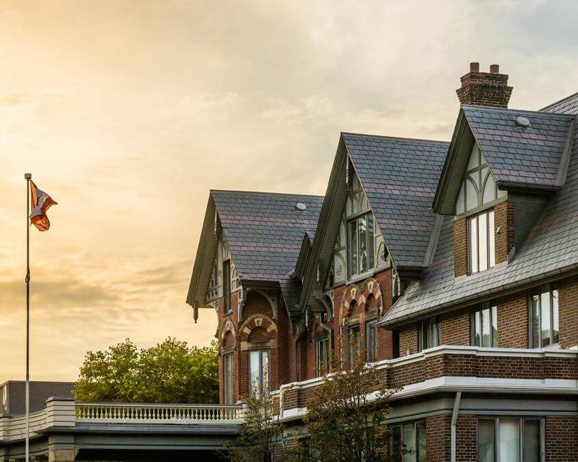 A historic brick building with gabled roofs and decorative trim stands under a cloudy sky at sunset. A Canadian flag on a tall pole is visible to the left. Trees and another building appear in the background.