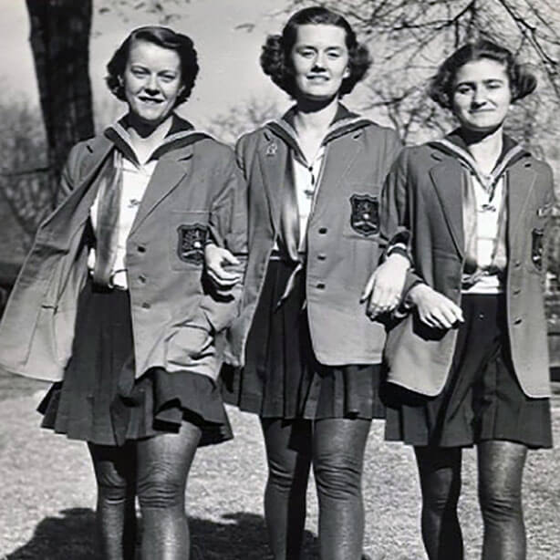 Three young women in matching jackets and pleated skirts walk arm-in-arm outdoors, smiling. The black-and-white photo suggests it was taken in the mid-20th century. Trees and grass are visible in the background.