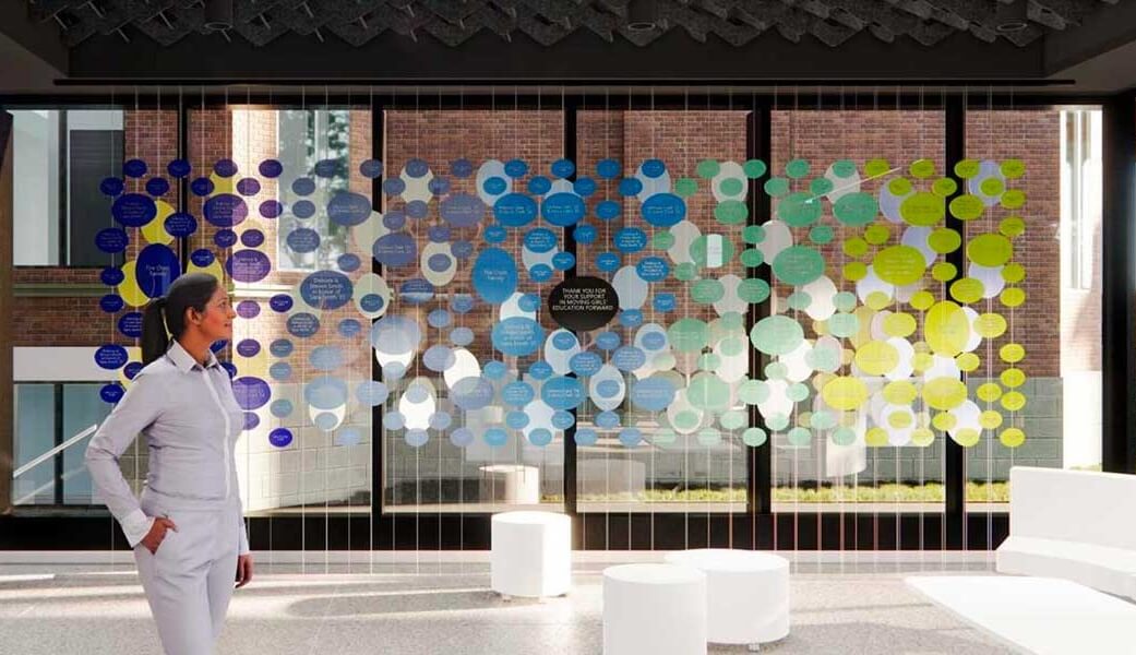 A person stands in a modern, bright room with large windows and colorful circles hanging in front of the glass, creating an artistic, abstract display. White modern furniture is arranged around the space.