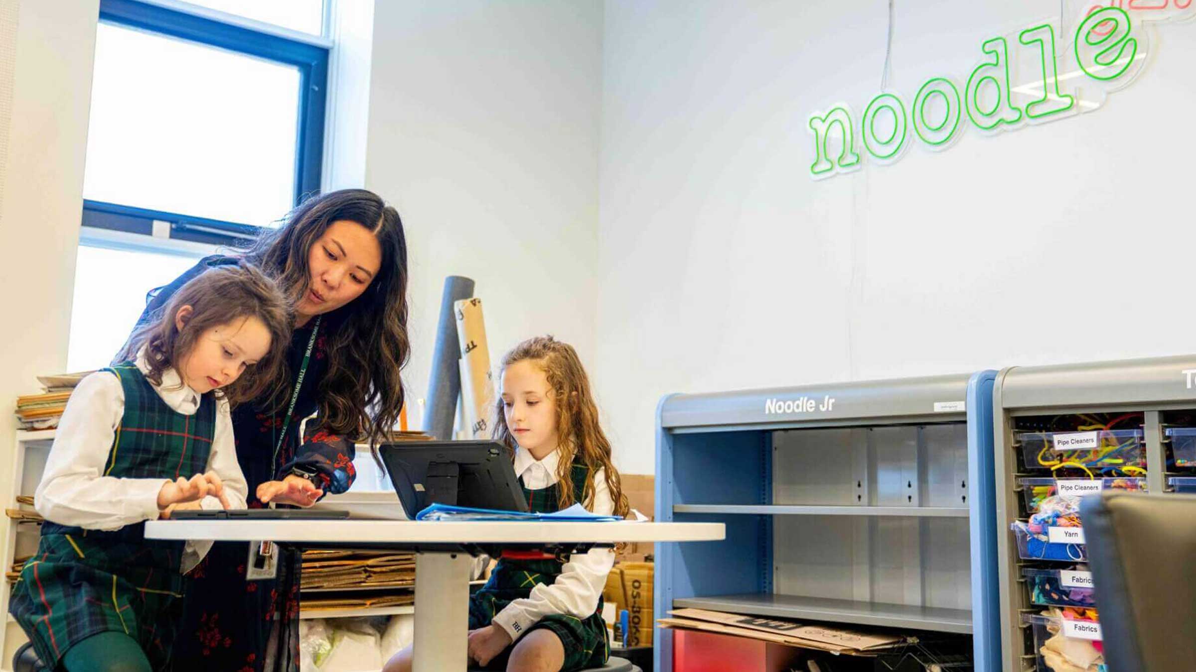 A teacher helps two young girls in school uniforms with a tablet at a classroom table. Shelves with educational materials are in the background, along with a neon "noodle" sign on the wall.