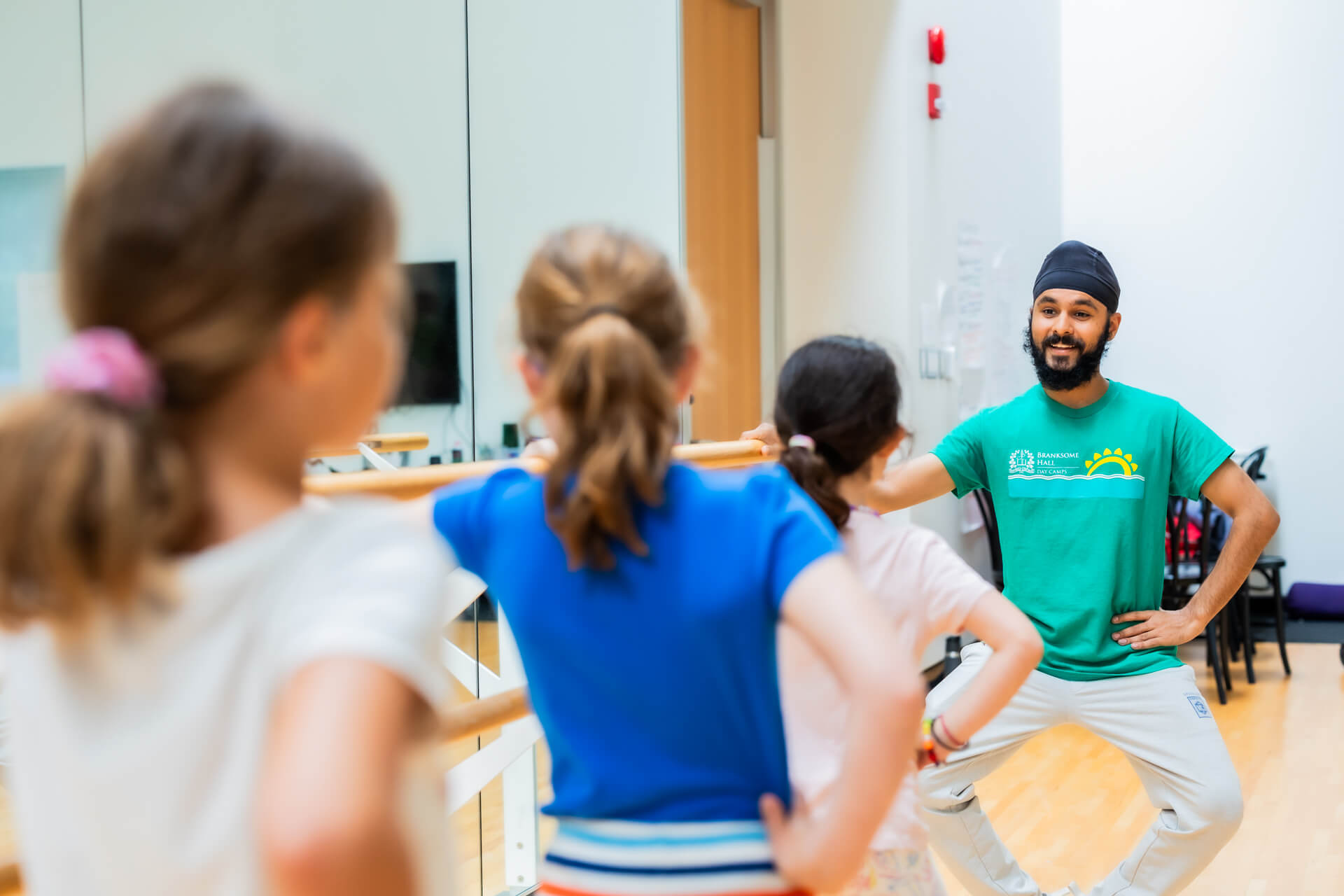 A dance instructor in a green shirt leads three young girls in a dance class; they stand at a barre with hands on hips, mirroring his pose in a brightly lit studio.