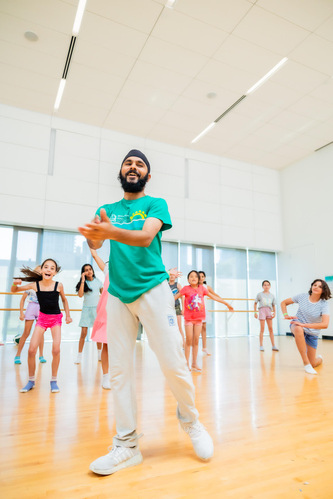 A man in a green shirt and white pants leads a dance class with enthusiastic children in a bright, modern studio with wooden floors and large windows.