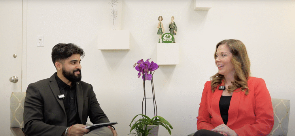A man in a black suit and a woman in a red blazer sit and talk in a bright room with white walls, a potted purple orchid between them, and decorative shelves with small figurines in the background.