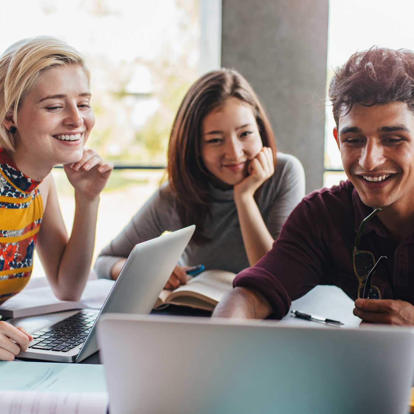 Three young adults sit together at a table, smiling and looking at laptops. Books and notebooks are open in front of them, suggesting they are studying or working on a group project in a bright, modern setting.