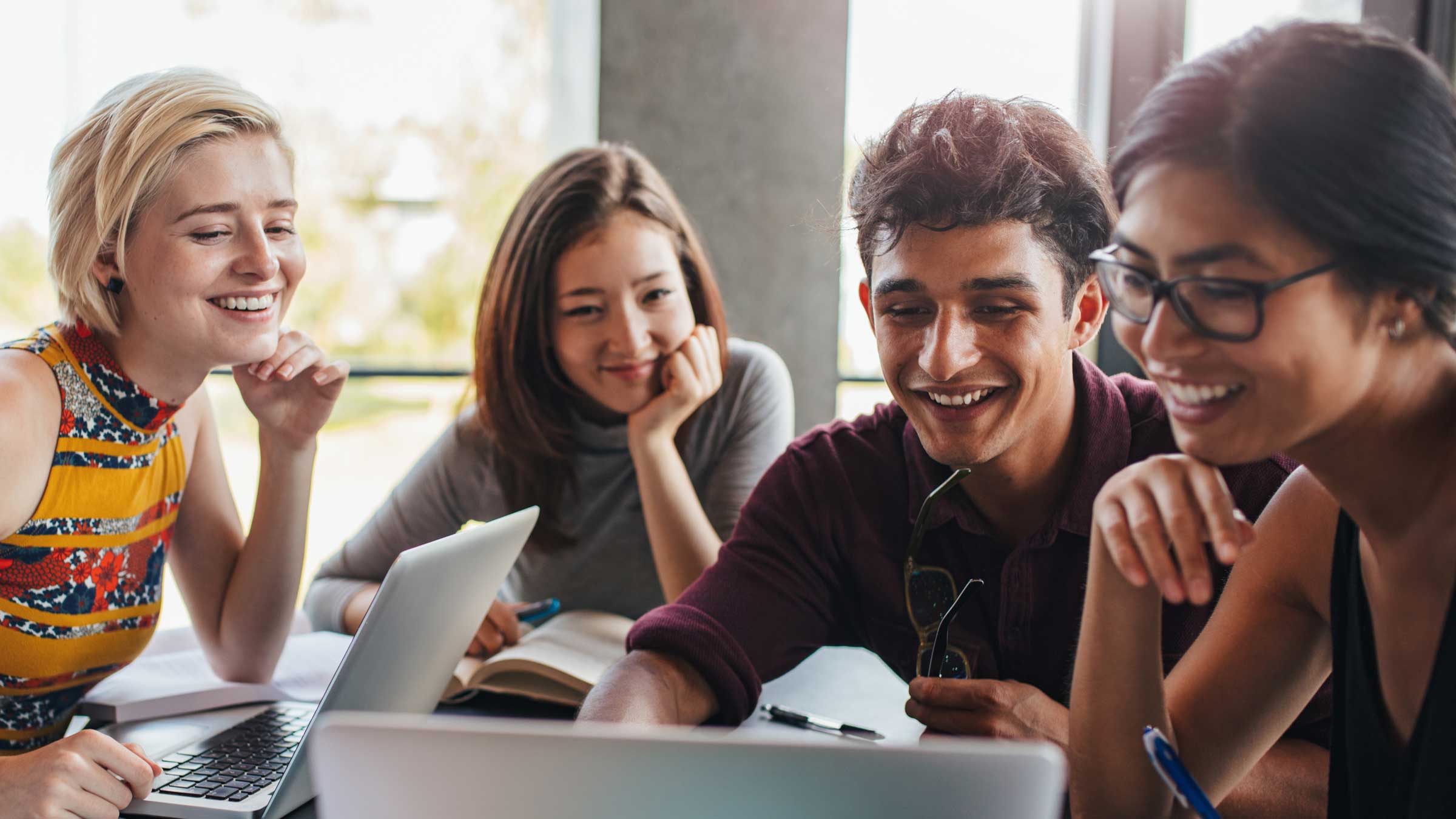 Four young adults sit together at a table, smiling and looking at a laptop screen. They appear to be collaborating or studying, with notebooks, pens, and another laptop on the table.
